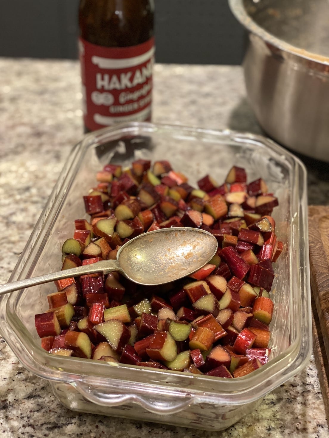 Diced rhubarb in a glass container with a spoon, next to a bottle of Hakanoa's syrup on a countertop.