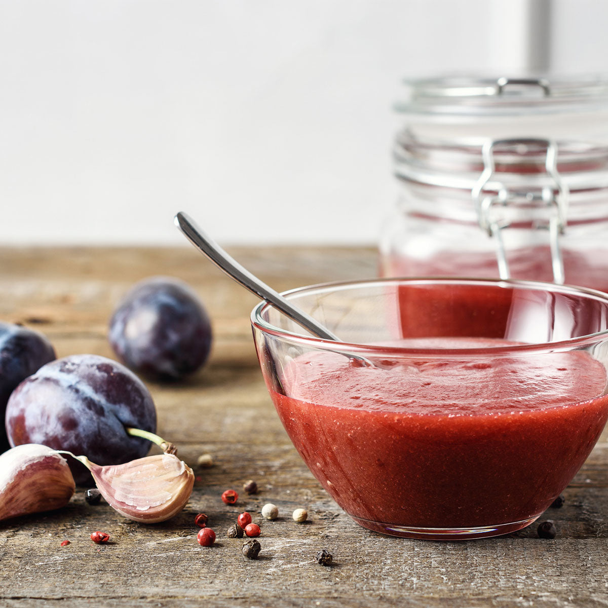 Glass bowl with red sauce on a wooden surface with plums and garlic cloves.