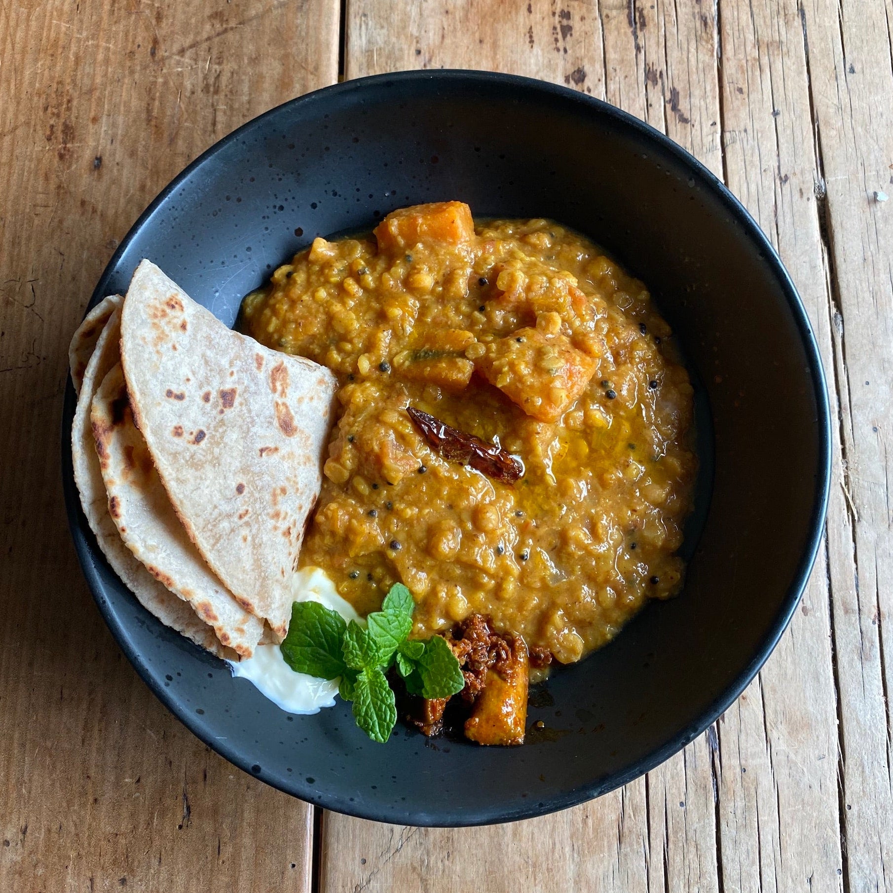 Black bowl with yellow lentil dhal soup, flatbread, and garnish on a wooden surface