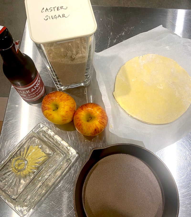 Ingredients and tools for baking on a metallic surface, including apples, flour, and a bottle of vanilla extract.